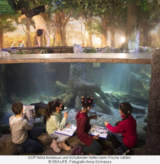 Der Artist springt ins Aquarium, die Kinder sitzen davor und zählen Fische