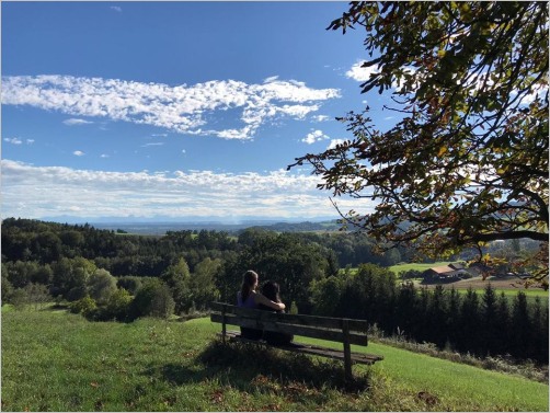 Frau mit Hund sitzt auf einer Bank mit wunderschönem Blick auf die hügelige Landschaft